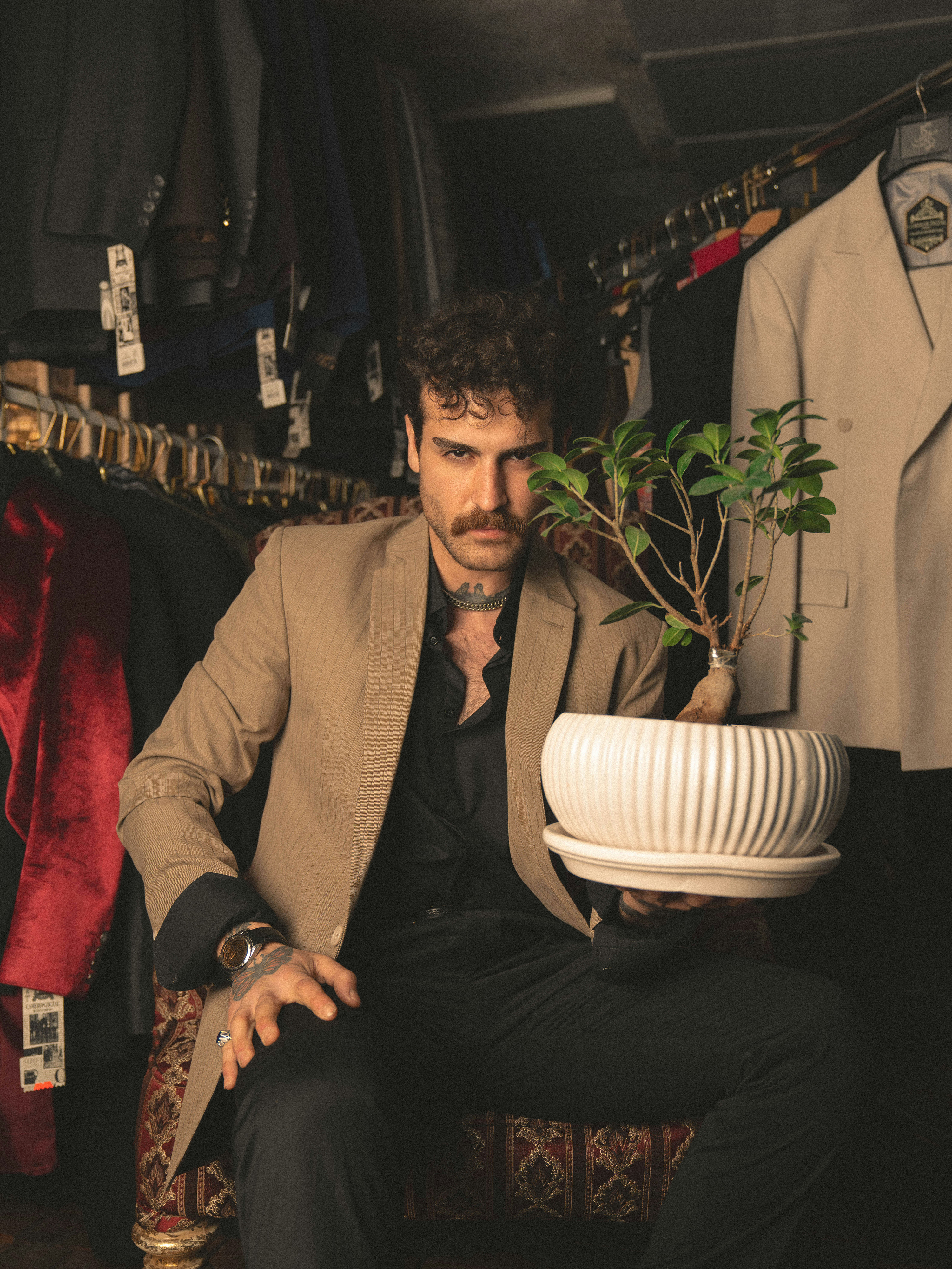 A man with a mustache sits in a clothing store holding a potted green plant in a white ribbed planter. He is wearing a beige blazer over a black shirt and black trousers.